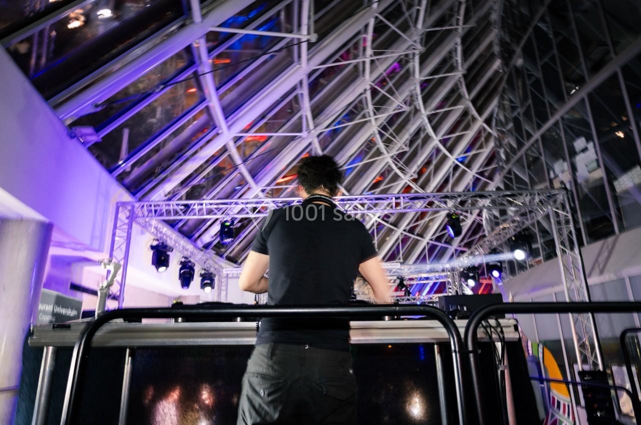 Un DJ de dos mixant sur une scène éclairée par des lumières colorées dans un espace moderne avec une verrière.