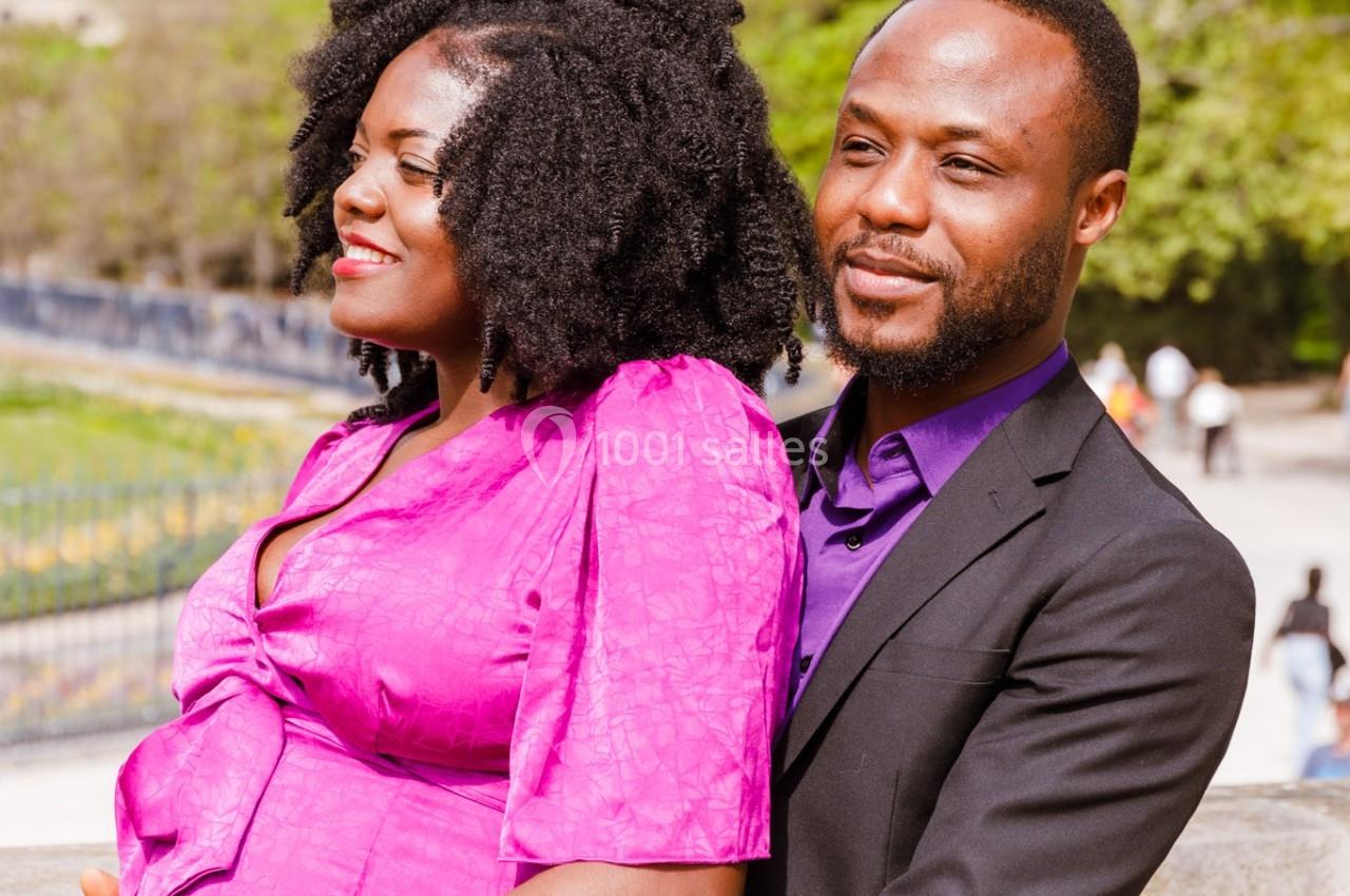 Un couple souriant pose sur un pont, avec un parc et des arbres en arrière-plan par temps clair.