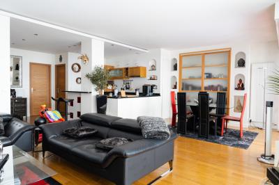 Femme assise sur un banc en extérieur, portant un sweat noir, avec un ciel bleu et des bâtiments en arrière-plan.