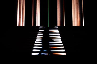 Femme assise sur un banc en extérieur, portant un sweat noir, avec un ciel bleu et des bâtiments en arrière-plan.