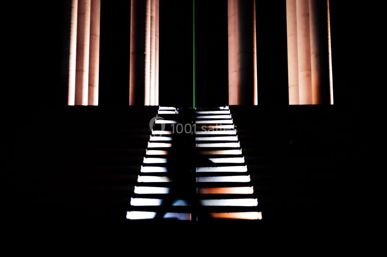 Silhouette floue d'une personne montant un escalier éclairé, entouré de colonnes lumineuses dans un environnement sombre.