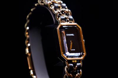 Femme assise sur un banc en extérieur, portant un sweat noir, avec un ciel bleu et des bâtiments en arrière-plan.