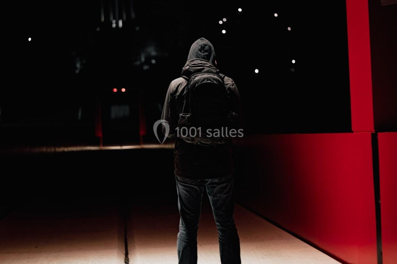 Un individu en manteau sombre et sac à dos, vu de dos, se tenant dans un espace faiblement éclairé avec des murs rouges.