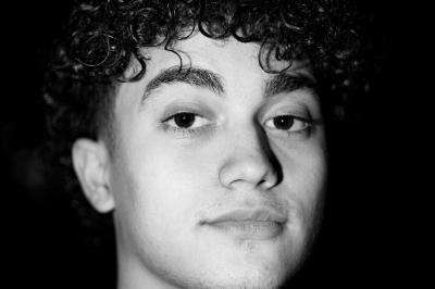 Femme assise sur un banc en extérieur, portant un sweat noir, avec un ciel bleu et des bâtiments en arrière-plan.