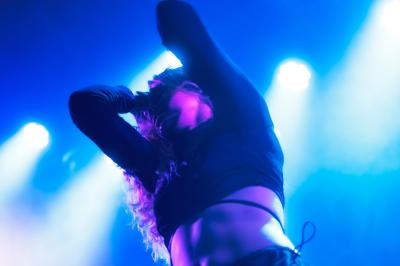 Femme assise sur un banc en extérieur, portant un sweat noir, avec un ciel bleu et des bâtiments en arrière-plan.