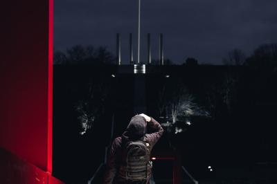 Femme assise sur un banc en extérieur, portant un sweat noir, avec un ciel bleu et des bâtiments en arrière-plan.