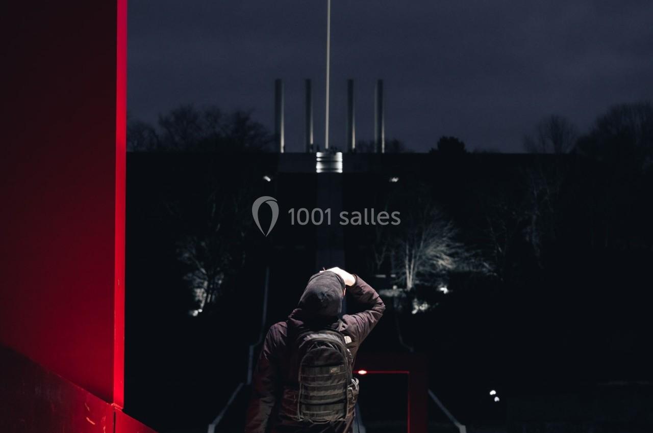 Un homme de dos avec un sac à dos observe un monument illuminé dans l'obscurité, entouré d'arbres.