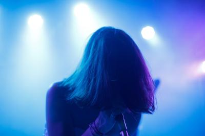 Femme assise sur un banc en extérieur, portant un sweat noir, avec un ciel bleu et des bâtiments en arrière-plan.