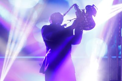 Femme assise sur un banc en extérieur, portant un sweat noir, avec un ciel bleu et des bâtiments en arrière-plan.
