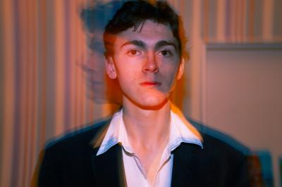 Femme assise sur un banc en extérieur, portant un sweat noir, avec un ciel bleu et des bâtiments en arrière-plan.