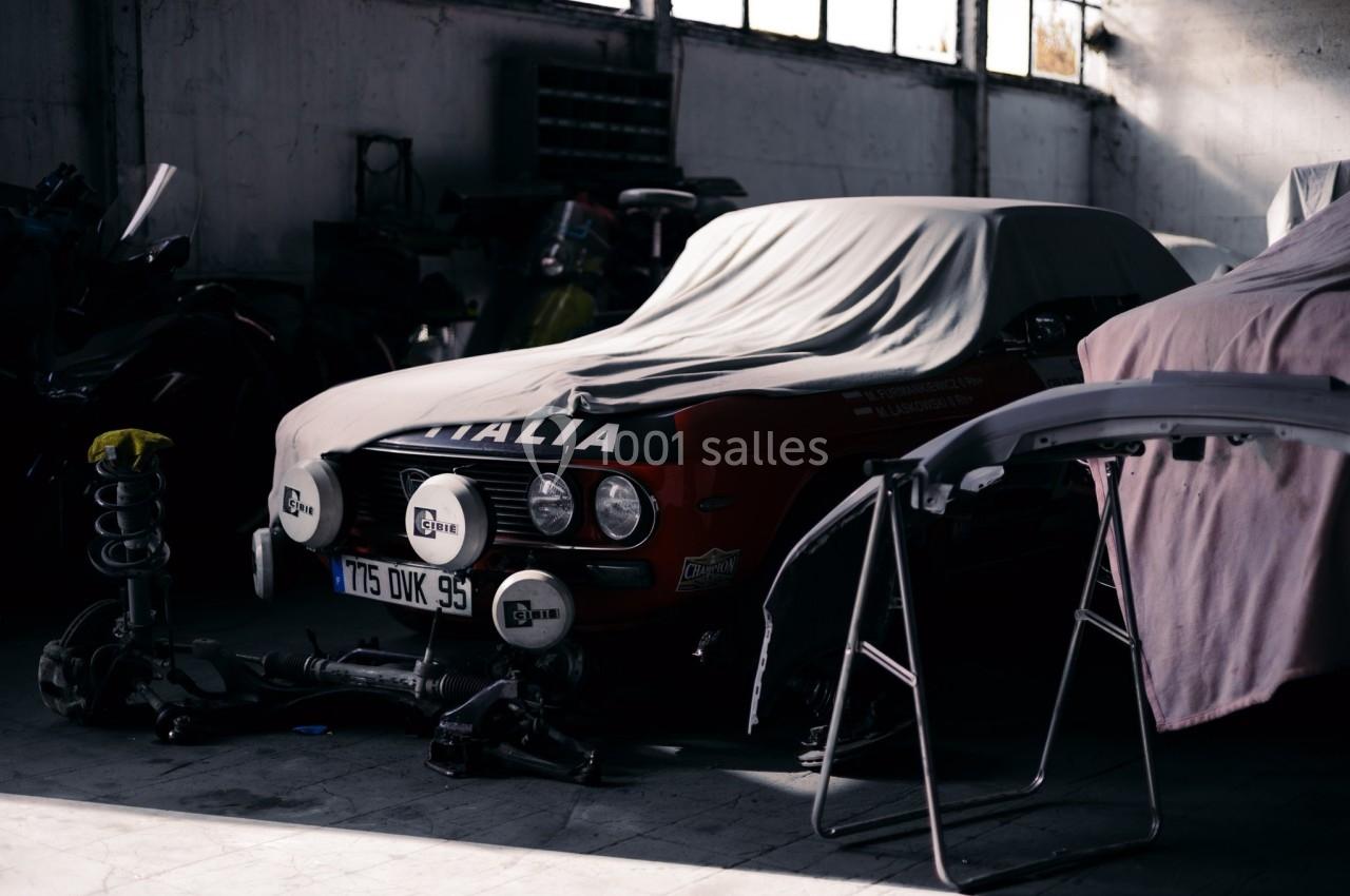 Voiture ancienne partiellement couverte dans un garage sombre, entourée de pièces détachées et d'outils.
