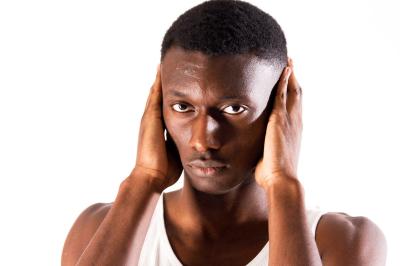 Femme assise sur un banc en extérieur, portant un sweat noir, avec un ciel bleu et des bâtiments en arrière-plan.