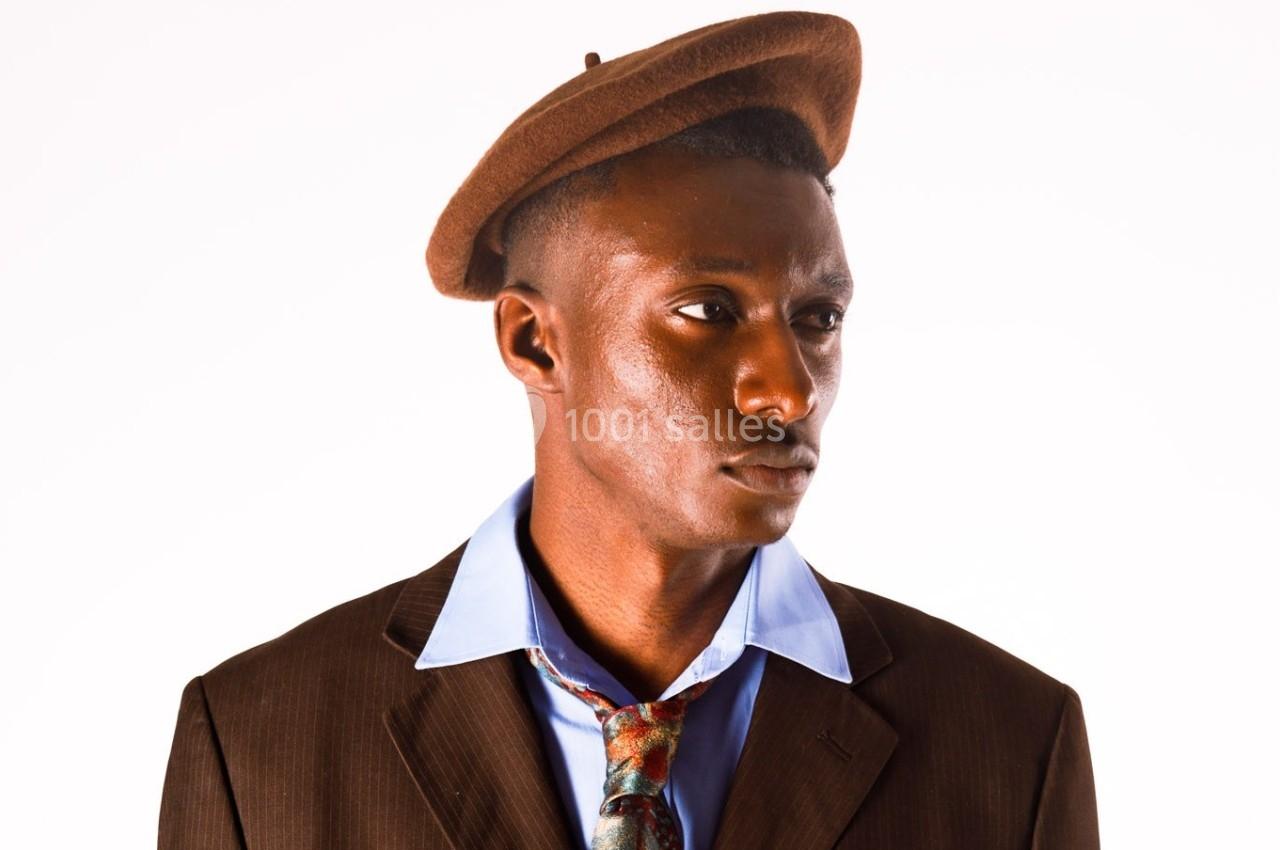 Homme portant un béret marron, une chemise bleue et une veste marron, avec une cravate légèrement dénouée.