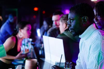 Femme assise sur un banc en extérieur, portant un sweat noir, avec un ciel bleu et des bâtiments en arrière-plan.