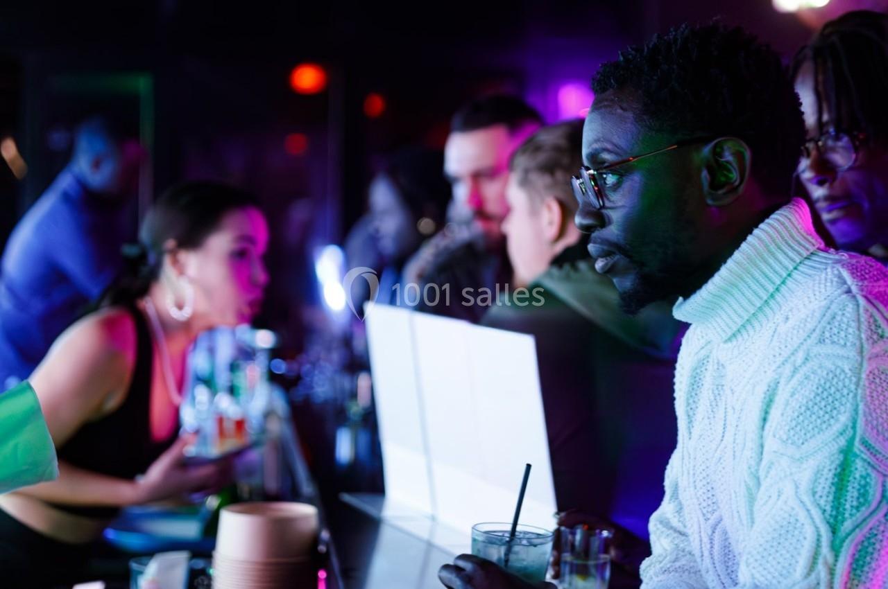 Un homme en pull blanc discute au comptoir d'un bar animé, entouré de personnes et de lumières colorées.