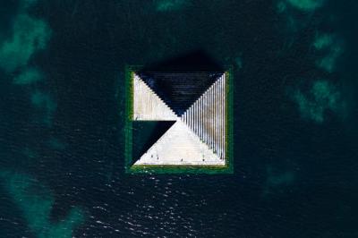 Femme assise sur un banc en extérieur, portant un sweat noir, avec un ciel bleu et des bâtiments en arrière-plan.