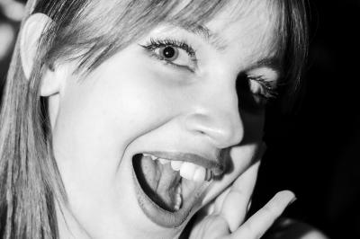 Femme assise sur un banc en extérieur, portant un sweat noir, avec un ciel bleu et des bâtiments en arrière-plan.