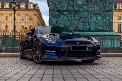 Femme assise sur un banc en extérieur, portant un sweat noir, avec un ciel bleu et des bâtiments en arrière-plan.