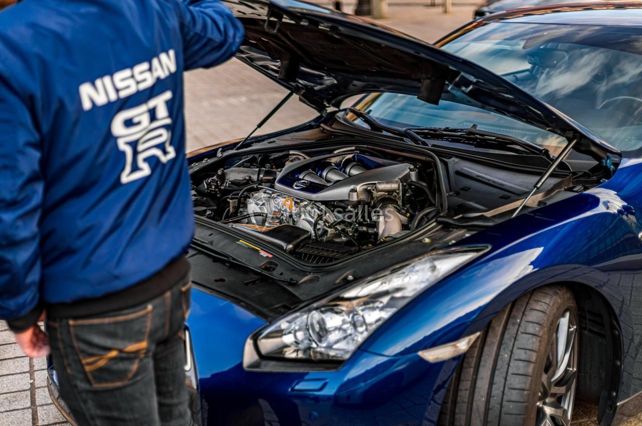 Un homme examine le moteur d'une voiture sportive bleue avec le capot ouvert, dans un environnement urbain.
