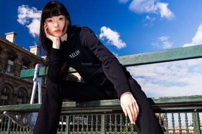 Femme assise sur un banc en extérieur, portant un sweat noir, avec un ciel bleu et des bâtiments en arrière-plan.
