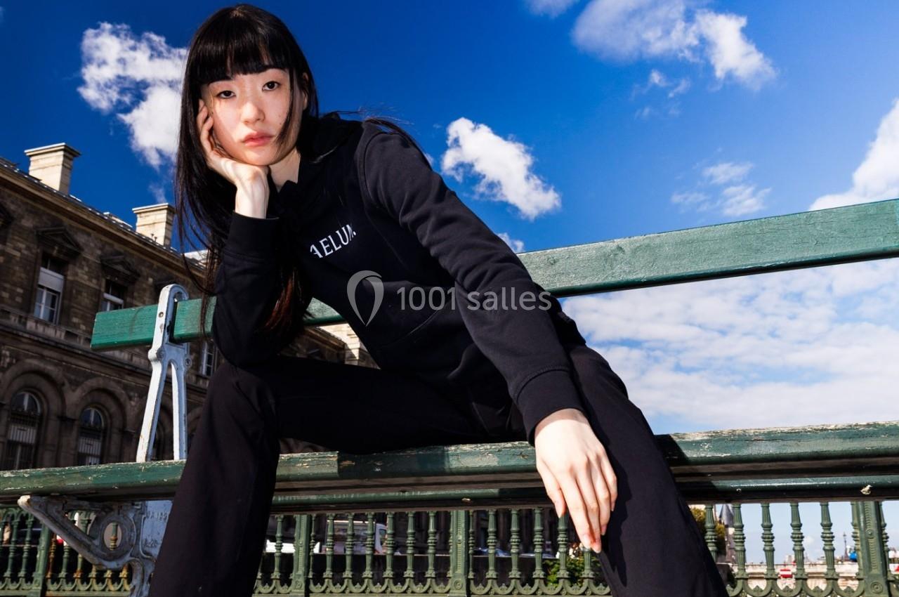 Femme assise sur un banc en extérieur, portant un sweat noir, avec un ciel bleu et des bâtiments en arrière-plan.