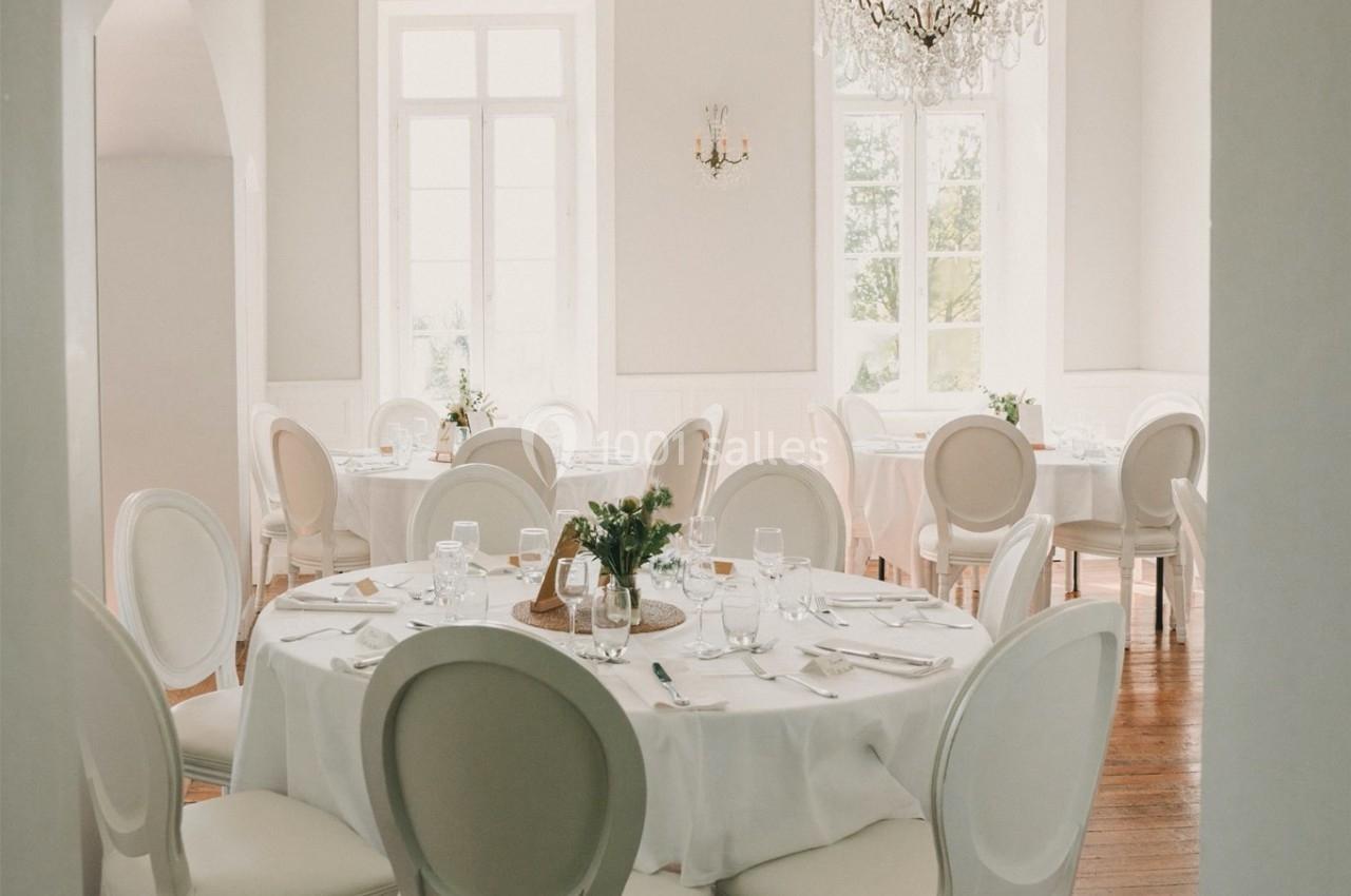 Salle lumineuse avec tables rondes dressées, chaises blanches élégantes, parquet en bois et lustre décoratif.