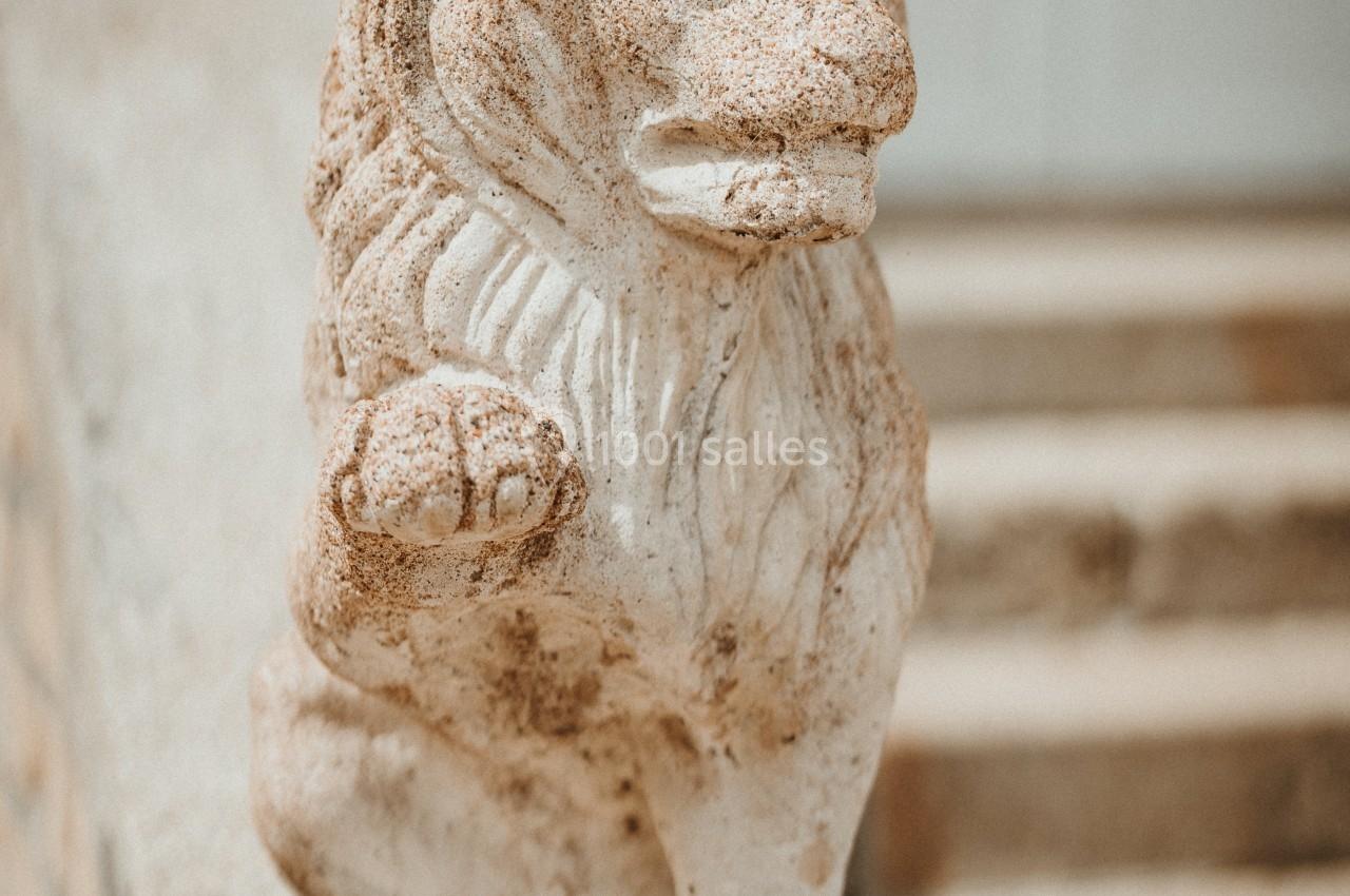 Statue en pierre d'un lion assis, posée sur un socle devant des marches en pierre et une porte blanche.