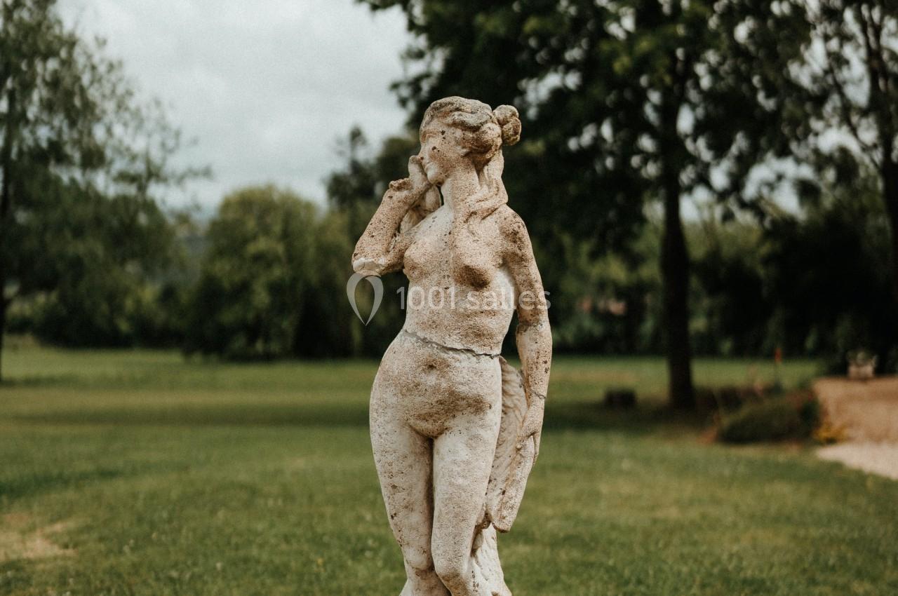 Statue en pierre représentant une femme nue, située dans un parc verdoyant sous un ciel partiellement nuageux.