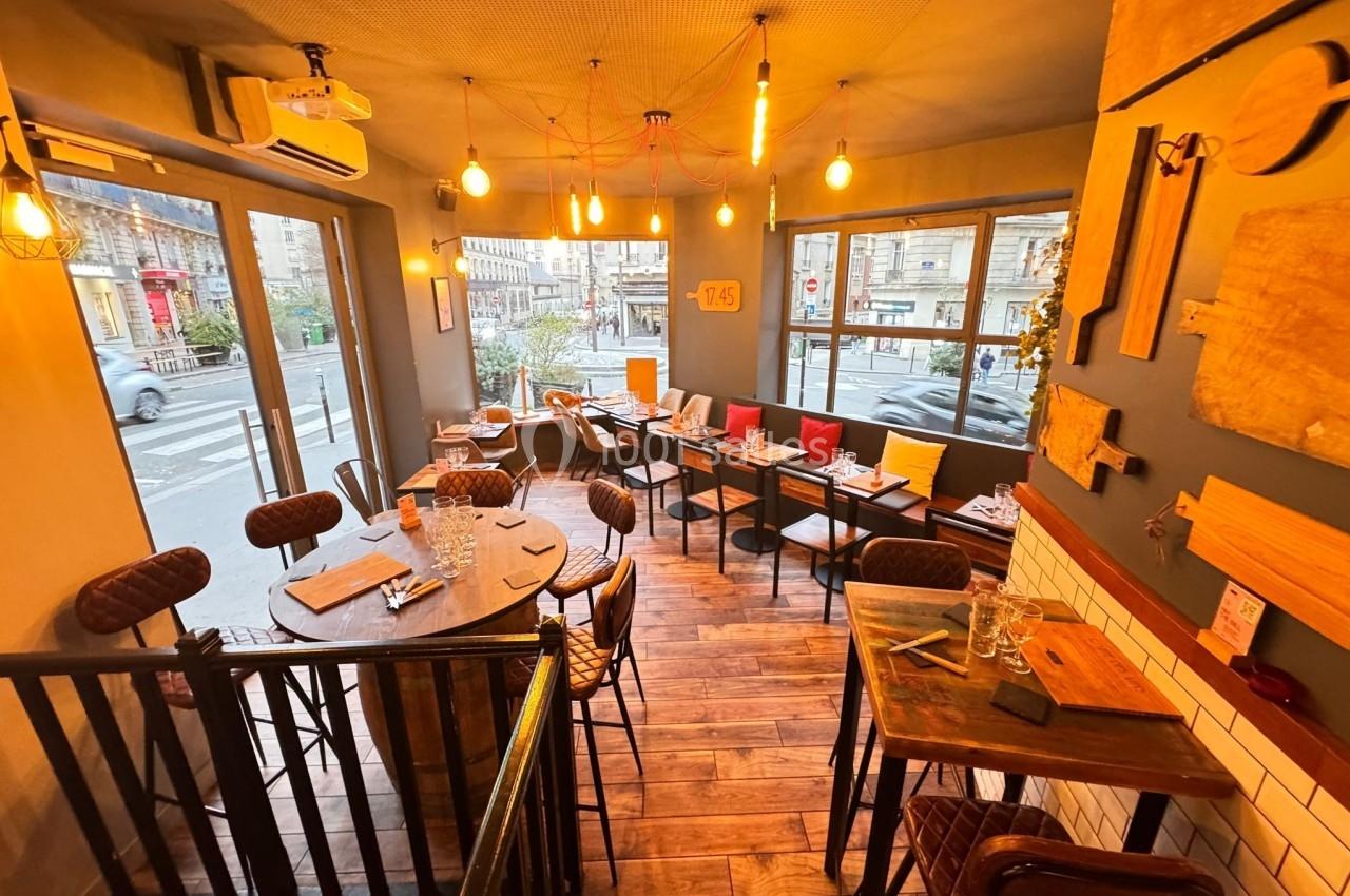 Salle de restaurant chaleureuse avec mobilier en bois, éclairage tamisé et grandes fenêtres donnant sur la rue.