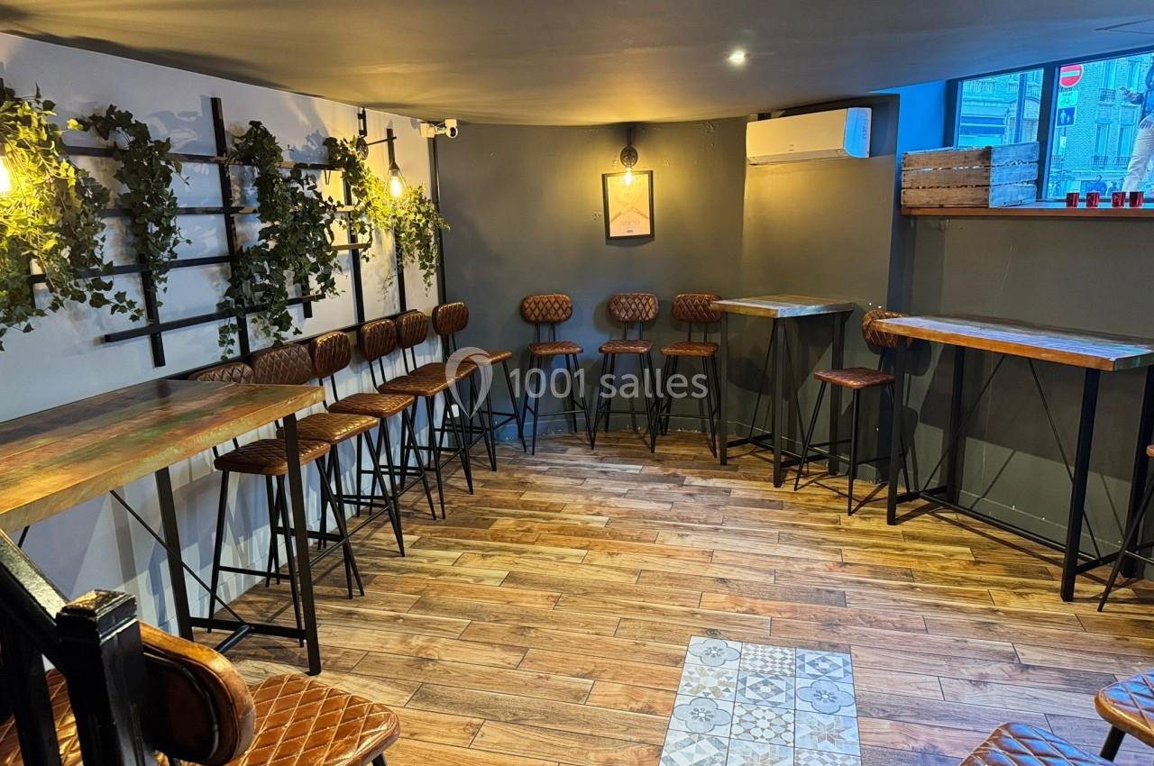 Salle avec tables hautes, tabourets en cuir, murs gris décorés de plantes et éclairage chaleureux.