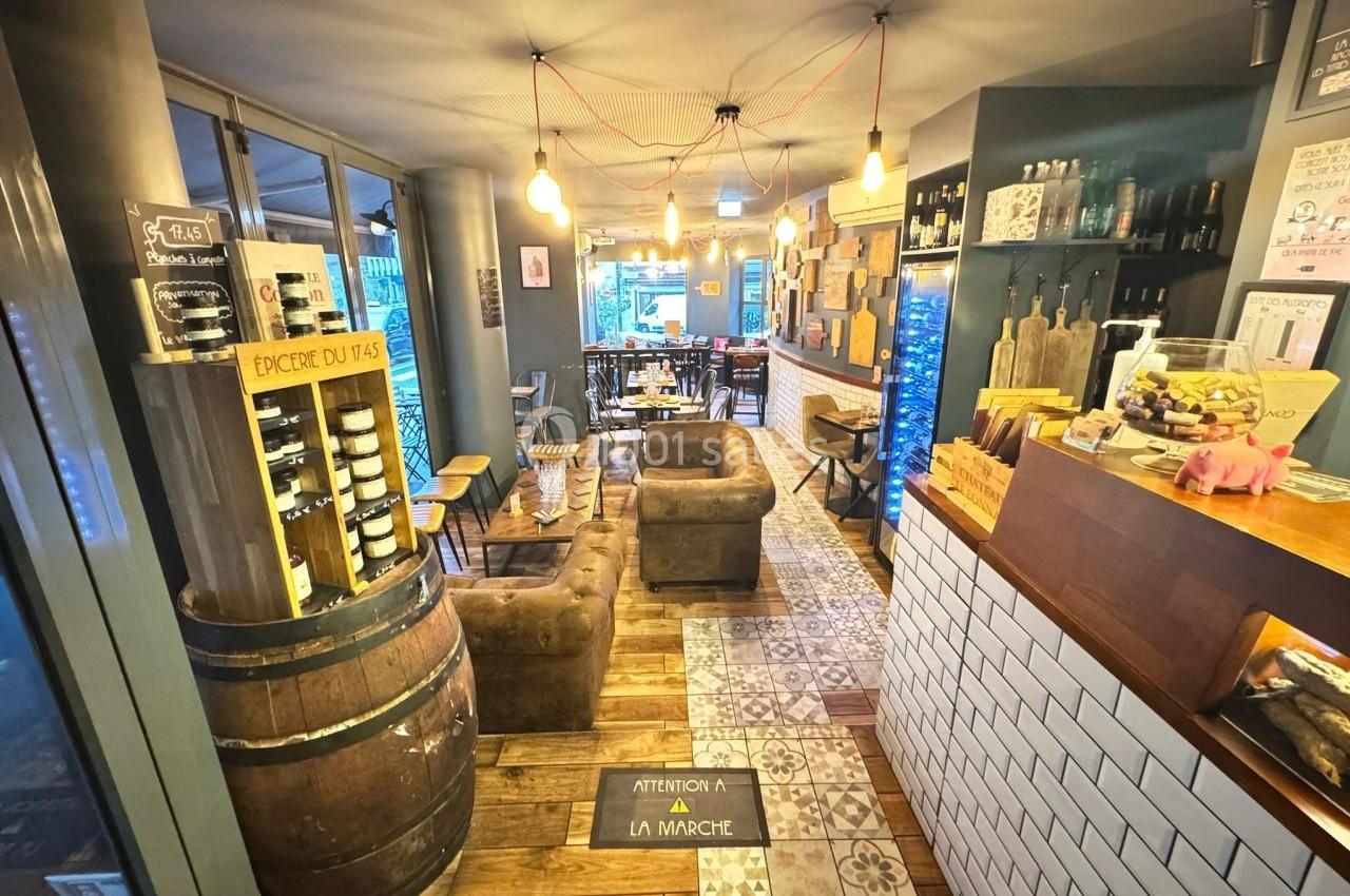 Intérieur d'un café chaleureux avec mobilier en bois, fauteuils, étagères de produits et comptoir en carrelage blanc.