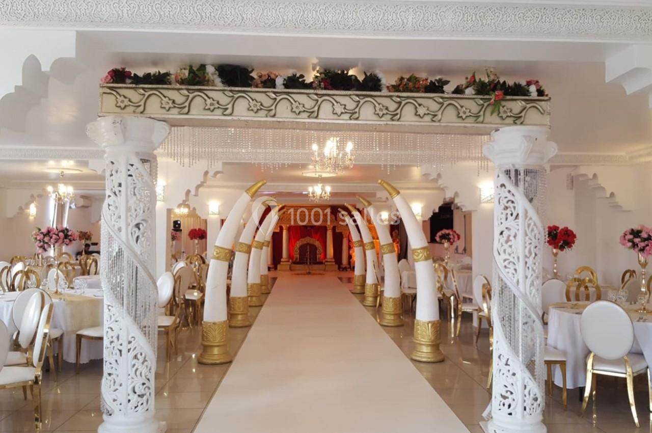 Salle de réception décorée avec colonnes sculptées, arches blanches et dorées, tables dressées et éclairage élégant.
