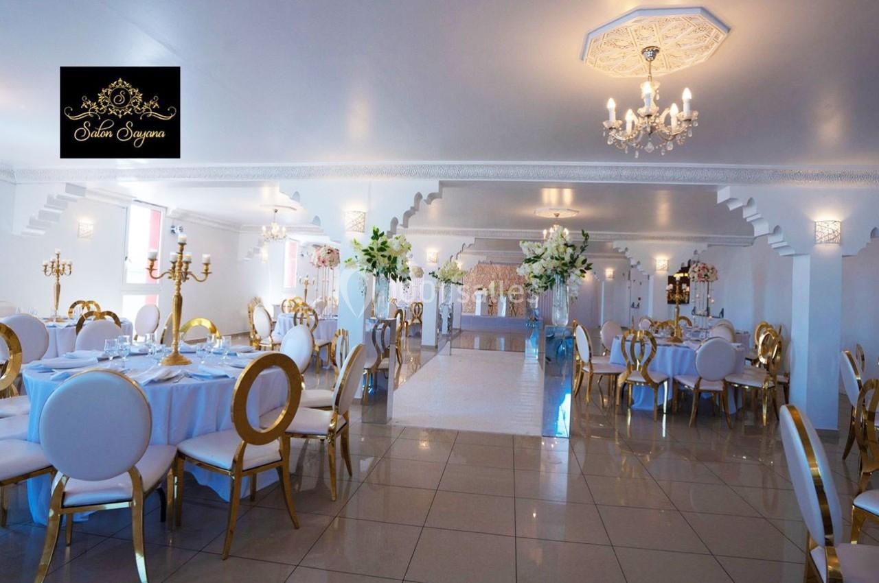 Salle de réception élégante avec tables dressées, chaises dorées et blanches, décor floral et lustres suspendus.