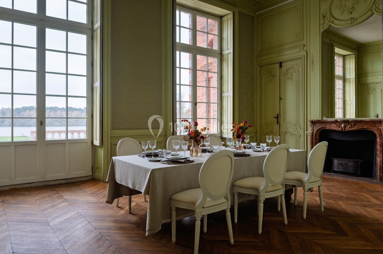 Salle à manger élégante avec table dressée, chaises blanches, cheminée en marbre et grandes fenêtres donnant sur l'extérieur.