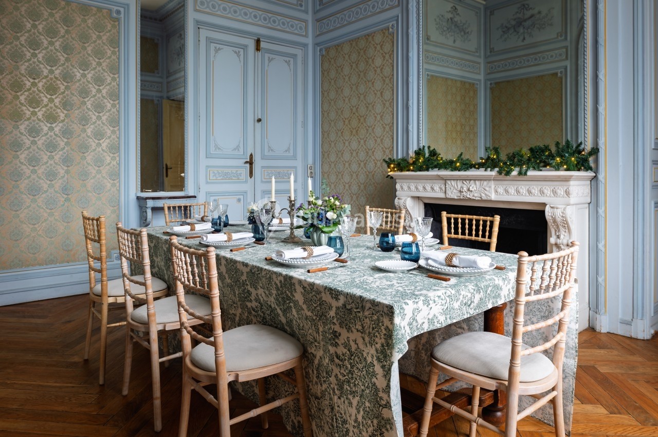 Table dressée avec une nappe à motifs, entourée de chaises en bois, dans une salle élégante avec cheminée décorée.