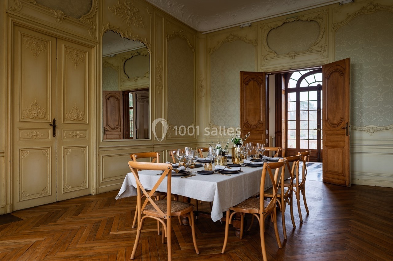 Salle à manger élégante avec table dressée, chaises en bois et portes-fenêtres ouvertes sur un parquet en chevrons.