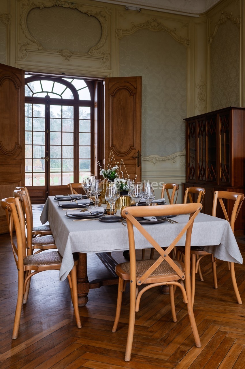Salle à manger élégante avec table dressée, chaises en bois, nappes blanches et grandes portes-fenêtres en arrière-plan.