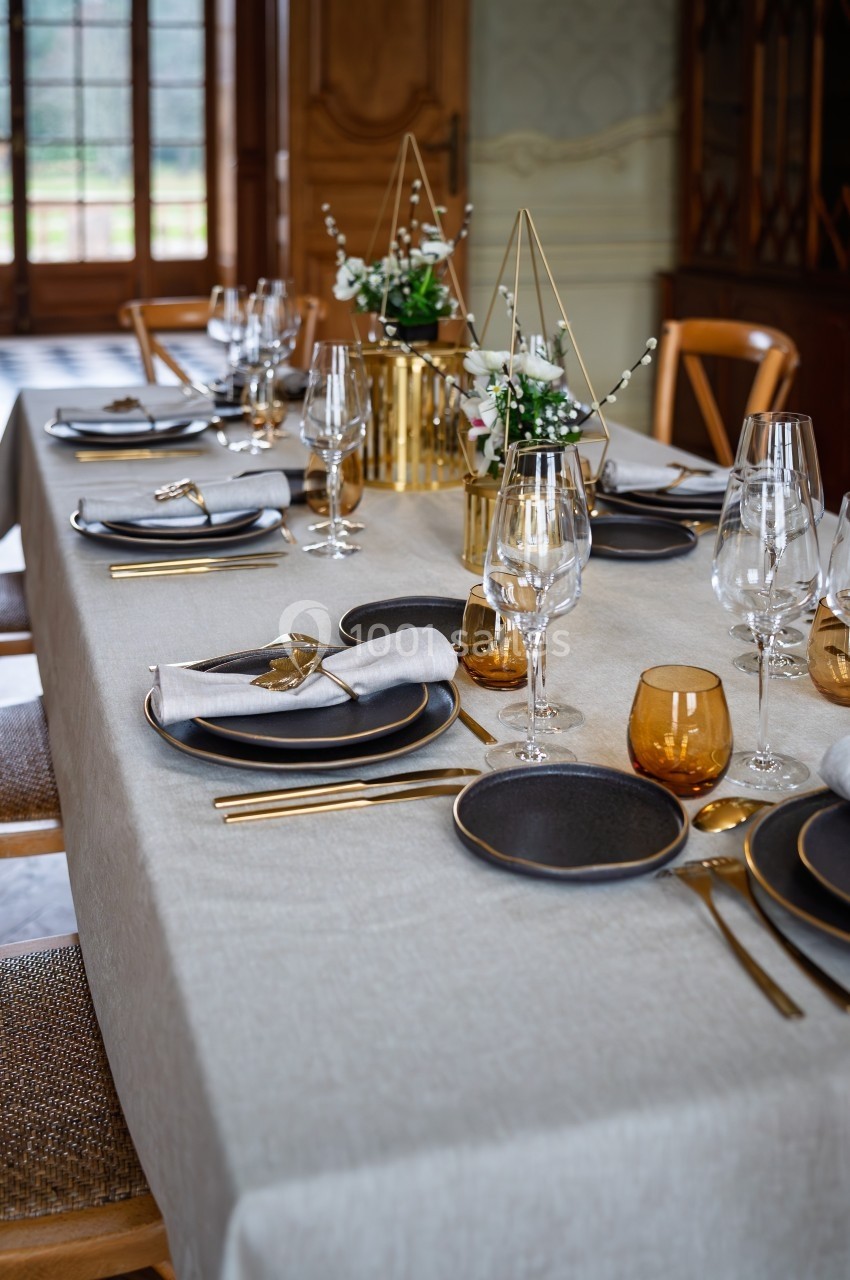 Table élégamment dressée avec vaisselle noire, couverts dorés, verres et décorations florales sur une nappe beige.