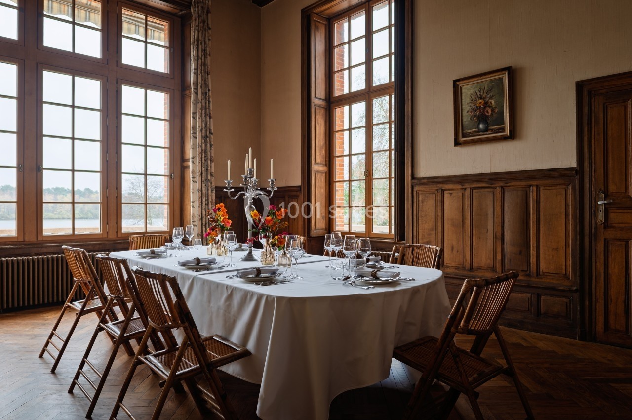 Table dressée avec nappes blanches, vaisselle et chandelier, entourée de chaises en bois dans une salle lumineuse au décor…