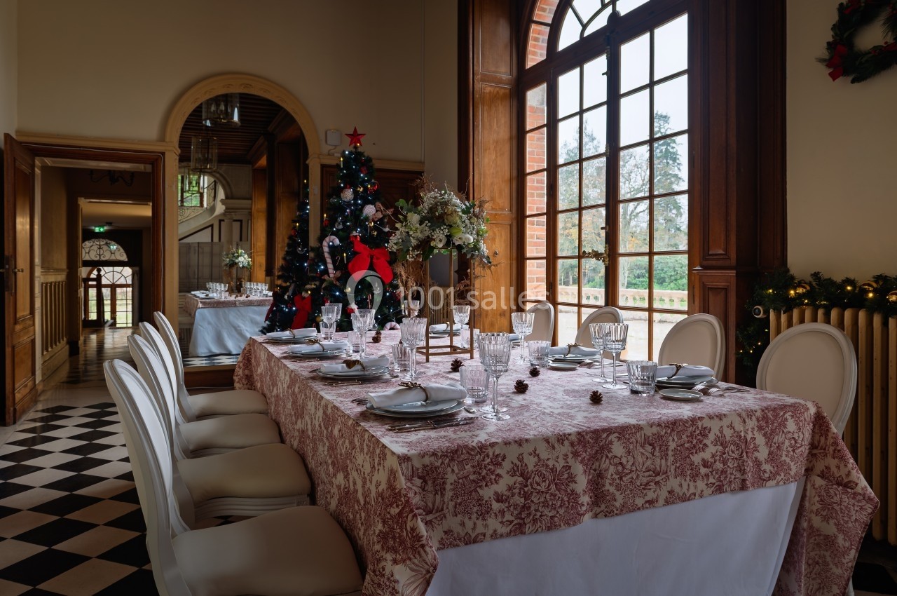 Salle à manger élégante avec table dressée, nappe à motifs, chaises blanches, sapin décoré et grande fenêtre donnant sur un…