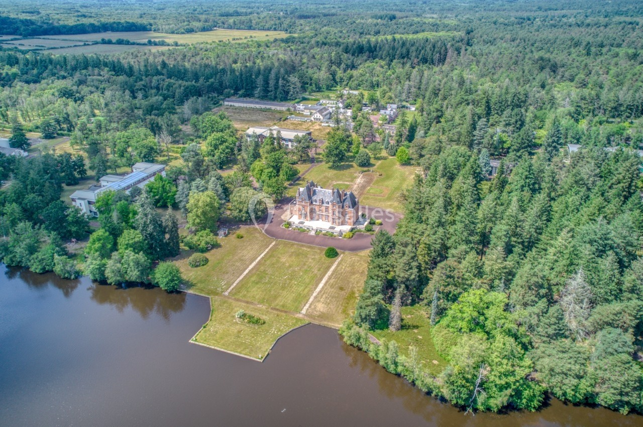 Vue aérienne d'un manoir entouré de forêt, avec un lac au premier plan et des bâtiments en arrière-plan.