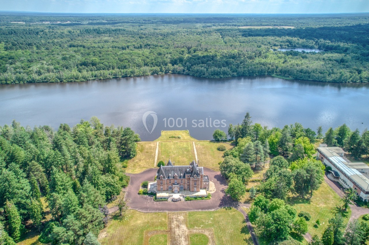 Vue aérienne d'un château entouré de forêt, situé au bord d'un grand lac sous un ciel dégagé.