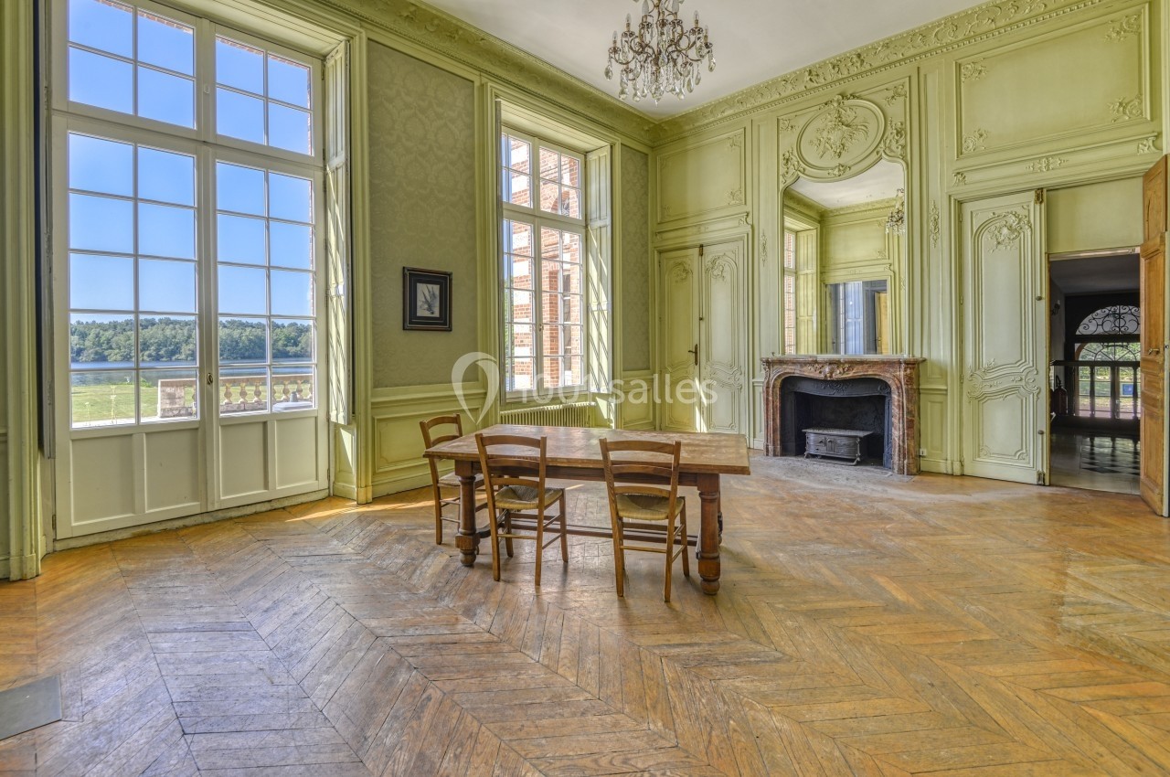 Salle lumineuse avec parquet en bois, cheminée décorative, murs ornés et vue sur un paysage extérieur verdoyant.