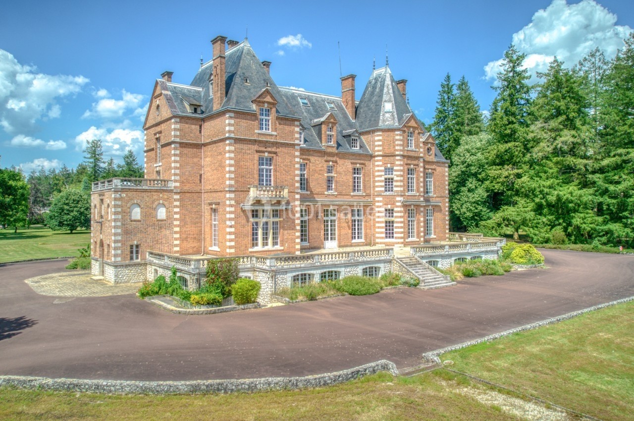 Château en briques rouges entouré de verdure, avec une allée pavée et un ciel dégagé en arrière-plan.