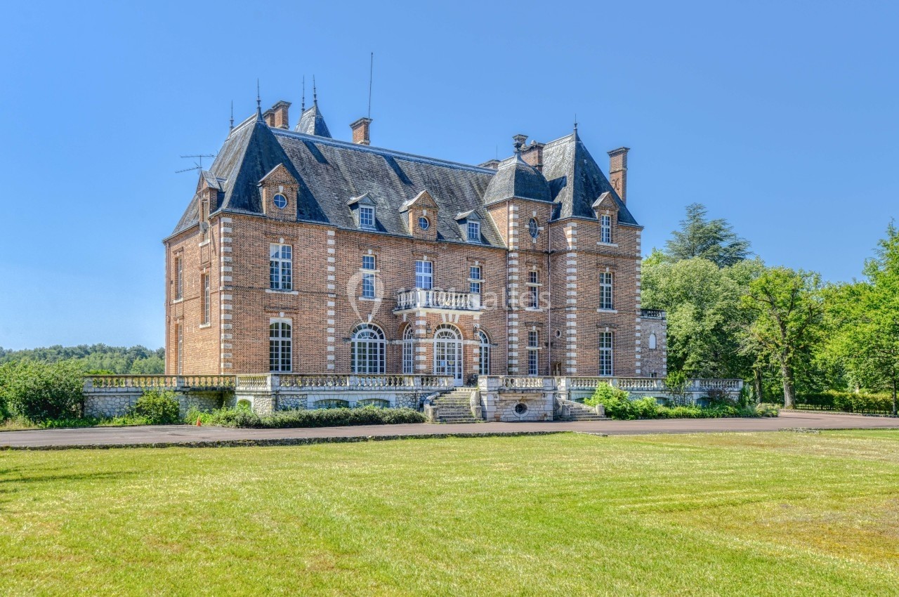 Façade d'un château en briques rouges avec toit en ardoise, entouré de verdure et d'une pelouse bien entretenue.