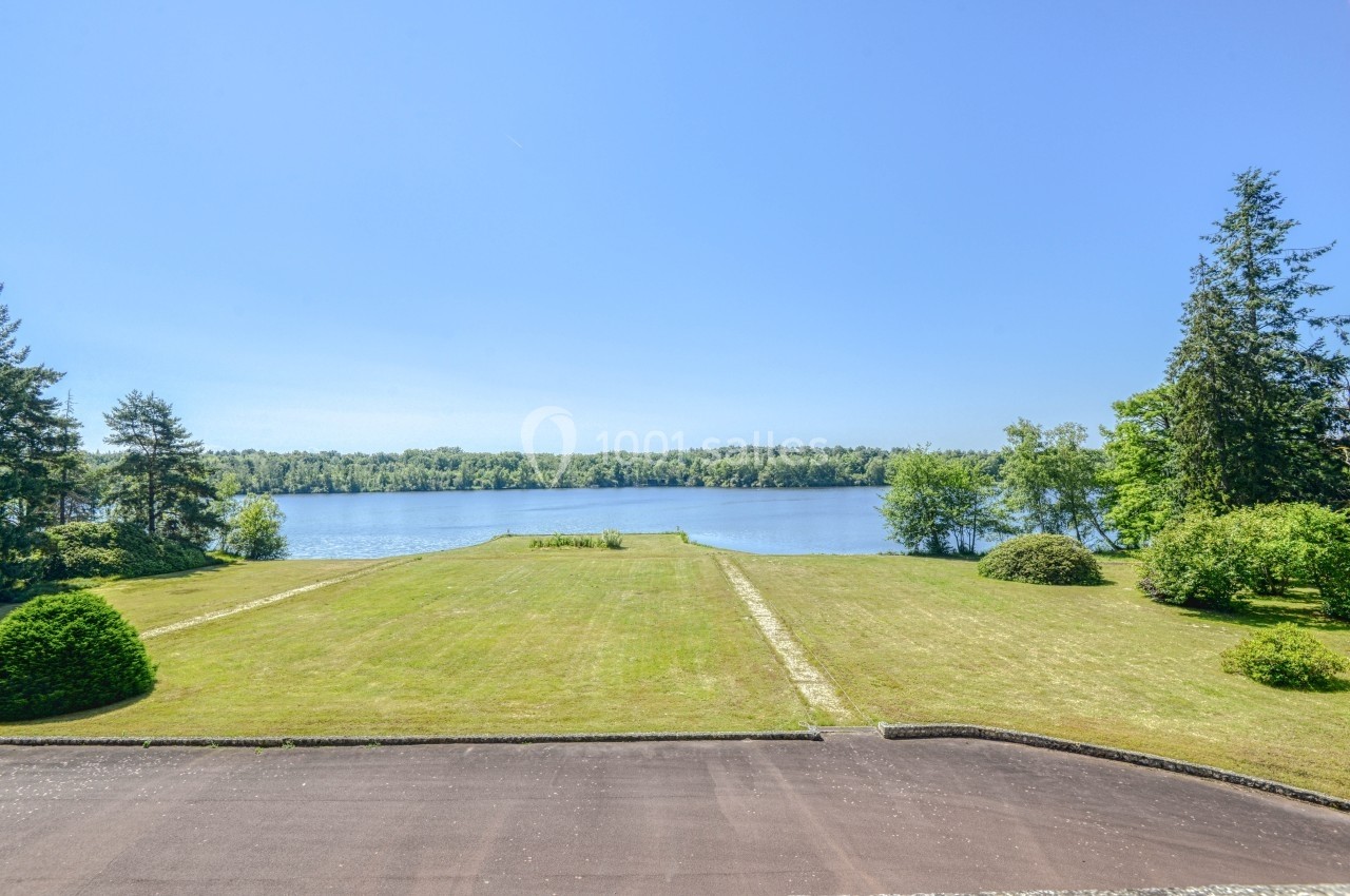 Vue sur un grand jardin verdoyant menant à un lac entouré d'arbres sous un ciel bleu dégagé.