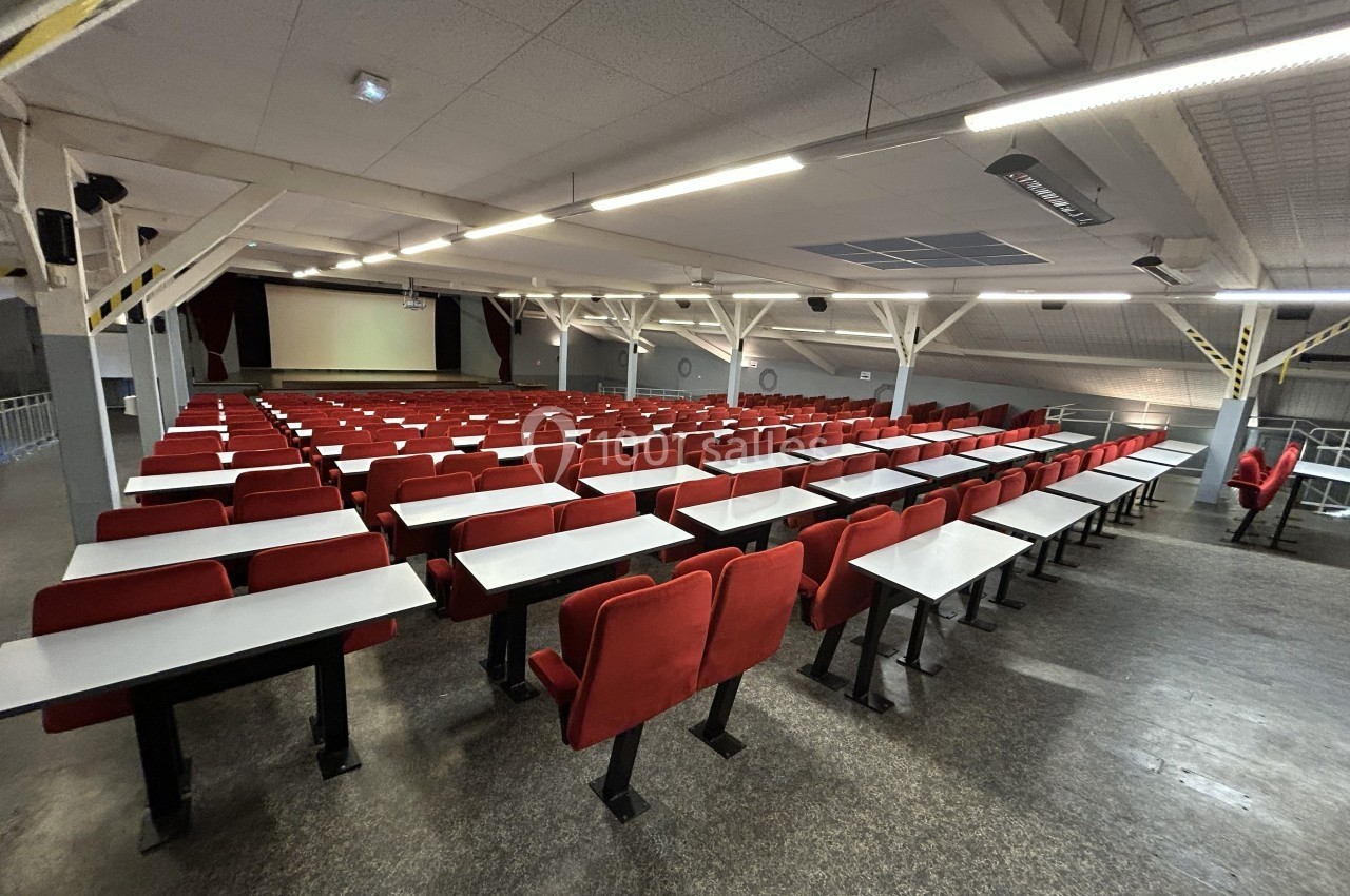 Salle de conférence vide avec rangées de sièges rouges et tables blanches, éclairée par des néons.