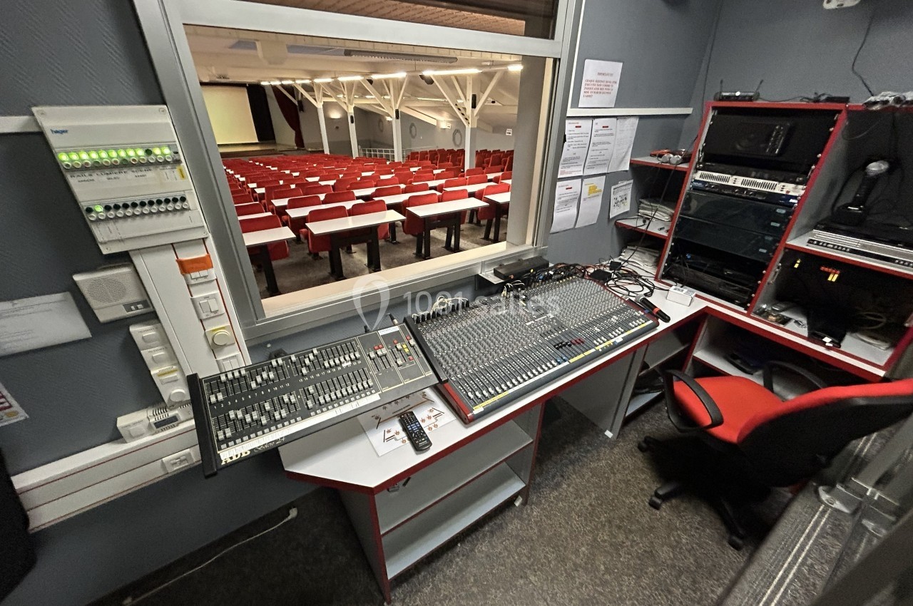 Salle de régie équipée de consoles de mixage audio et vidéo, avec vue sur une salle de conférence vide.