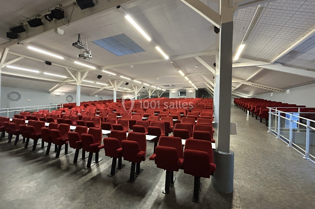Salle de conférence vide avec des rangées de sièges rouges, éclairée par des lumières au plafond.