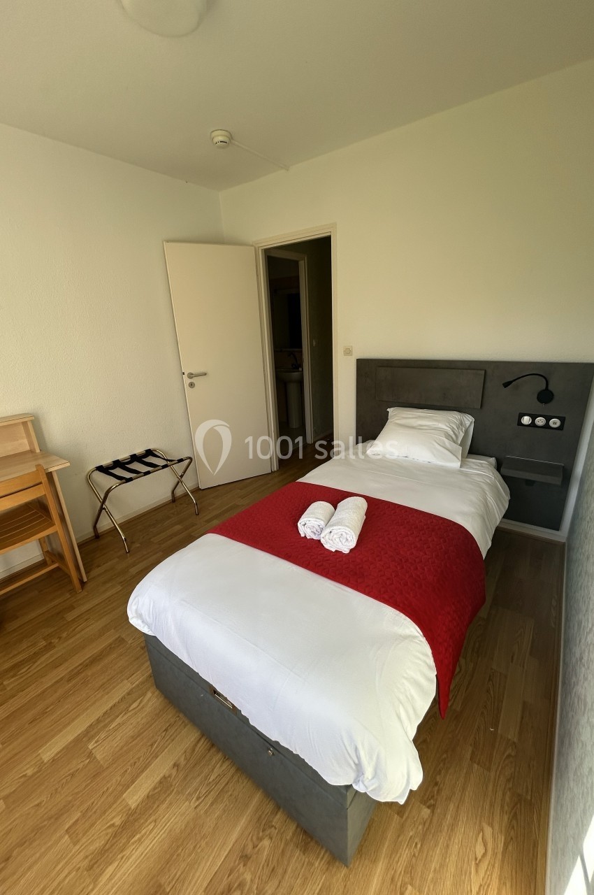 Chambre simple avec lit une place, drap rouge, bureau en bois, porte ouverte sur une autre pièce.